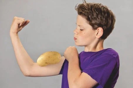 Young boy in a purple t-shirt flexing his arm against a grey background with a large potato placed on his bicep to humorously mimic a strong muscle.
