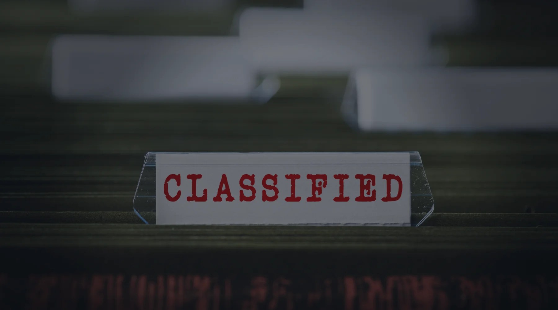 Close-up of a white label with red stamped CLASSIFIED text inside a clear plastic folder tab, positioned in a dark office filing drawer with blurred background folders.