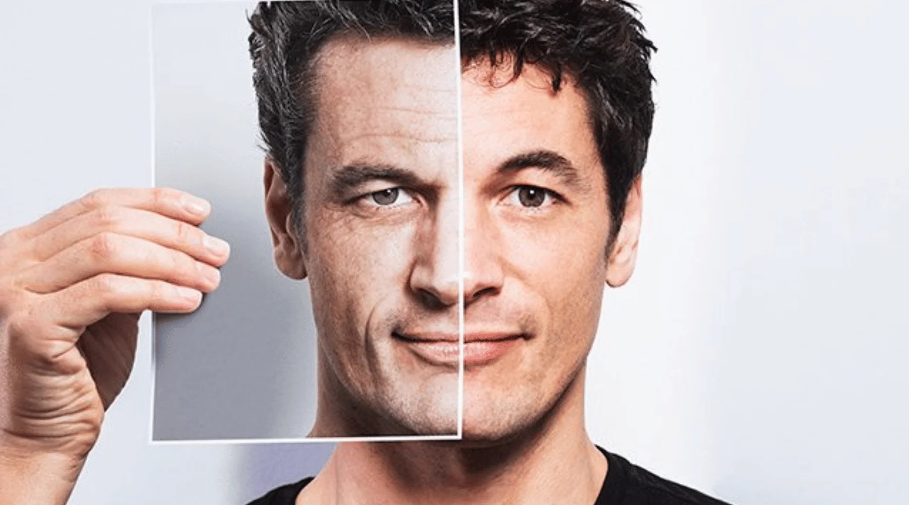 Man holding a printed photograph of his older, wrinkled face over his younger, smoother face against a plain background, illustrating the effects of aging and skin rejuvenation treatments.