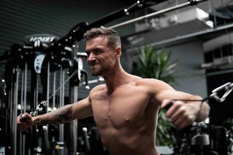 Shirtless muscular man with an arm tattoo performs cable chest flies on a Hoist machine in a gym, showing defined pectoral muscles and focused effort against a dark, blurred background.