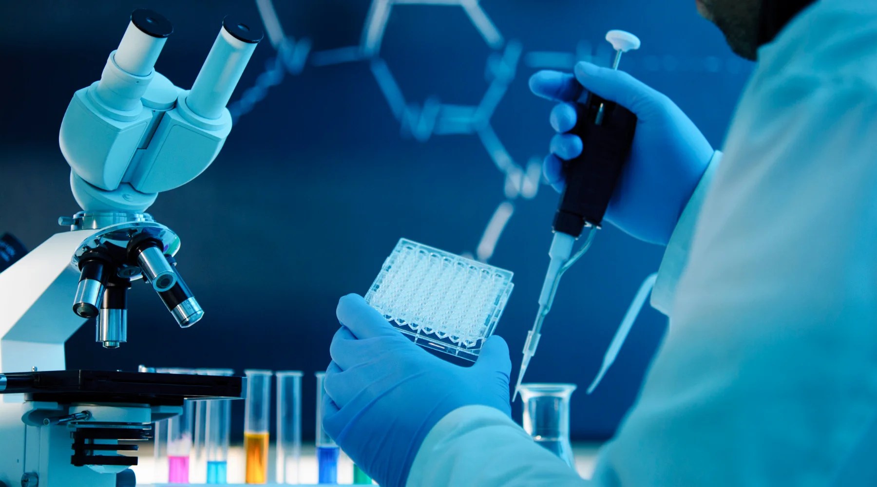 Researcher in blue gloves holding a multi-well plate and using a pipette in a laboratory with a microscope and colorful test tubes against a blue chemical structure background.