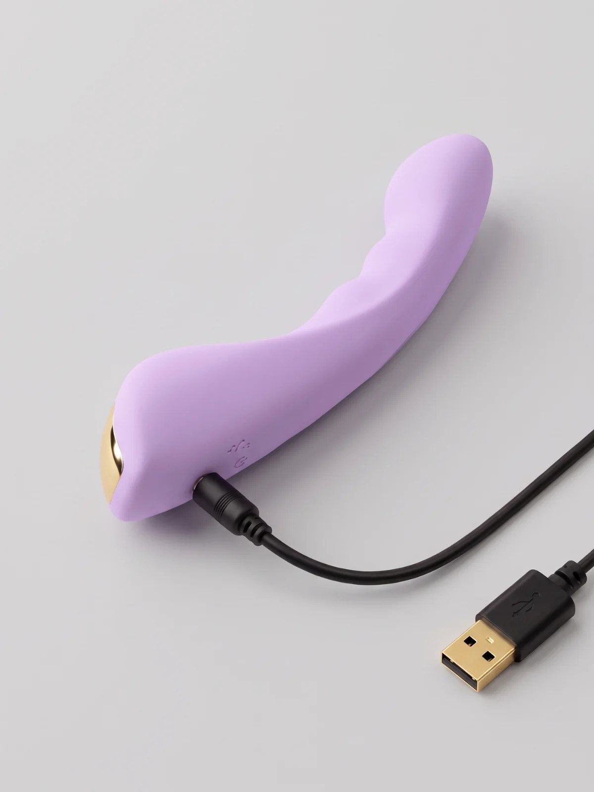 Vesper rechargeable lavender G-spot vibrator with a curved tip and gold accent, connected to its USB charging cable.
