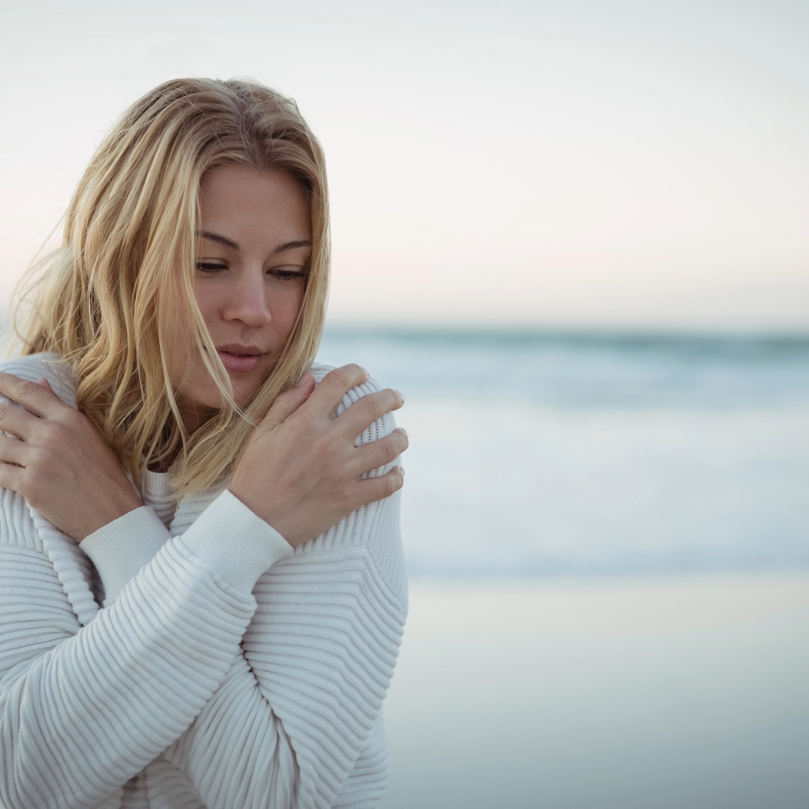Woman in white sweater embracing herself on a serene beach, symbolizing emotional healing and self-compassion in somatic therapy