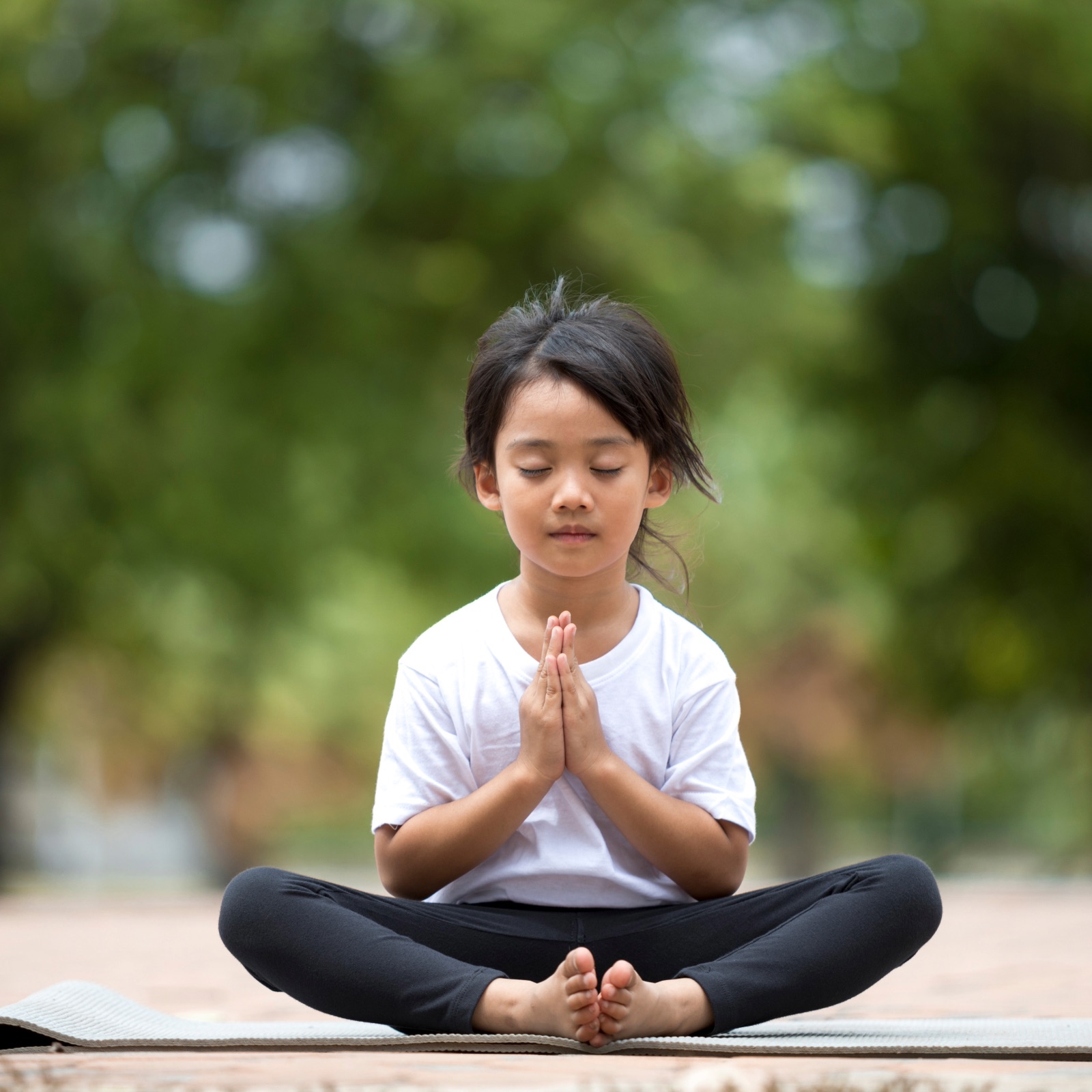 A young Asian girl in a white t-shirt and black leggings sits cross-legged on a yoga mat, practicing meditation in a soft-focused green, natural outdoor setting. Her eyes are closed, and her hands are pressed together in a prayer position, embodying calm and inner peace.