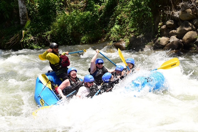 A group of people wearing helmets and life jackets whitewater rafting through rushing rapids in a river, paddling together in an inflatable raft surrounded by lush green jungle scenery.