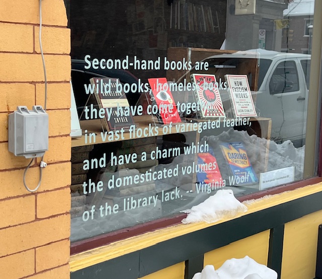 Bookstore window display at City Books in Pittsburgh featuring a quote about second-hand books, with stacks of used books visible inside and snow resting along the window ledge.