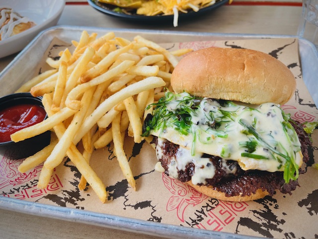 Open-faced cheeseburger topped with melted cheese and fresh herbs, served alongside a pile of golden French fries and a small cup of ketchup on a paper-lined tray at a casual restaurant.