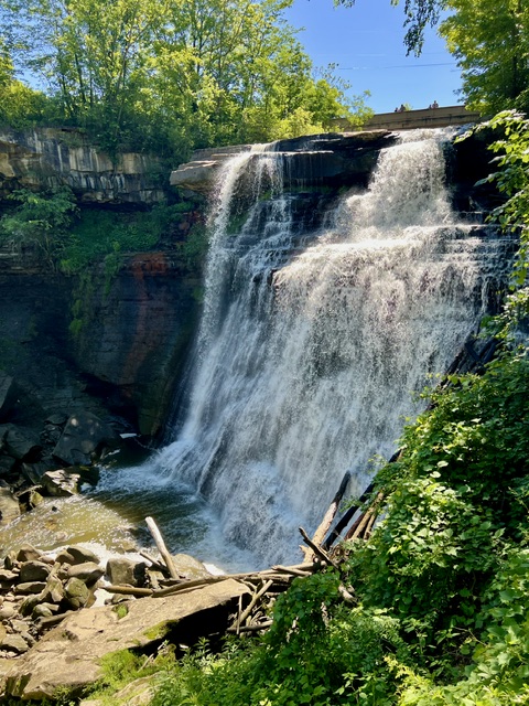 Brandywine Falls cascading over a layered sandstone cliff in Cuyahoga Valley National Park, surrounded by lush green trees and vegetation on a bright summer day.