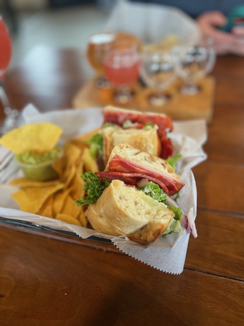 Italian-style sub sandwich served with kettle chips and tortilla chips with guacamole at a local brewery, with a flight of craft beers blurred in the background.
