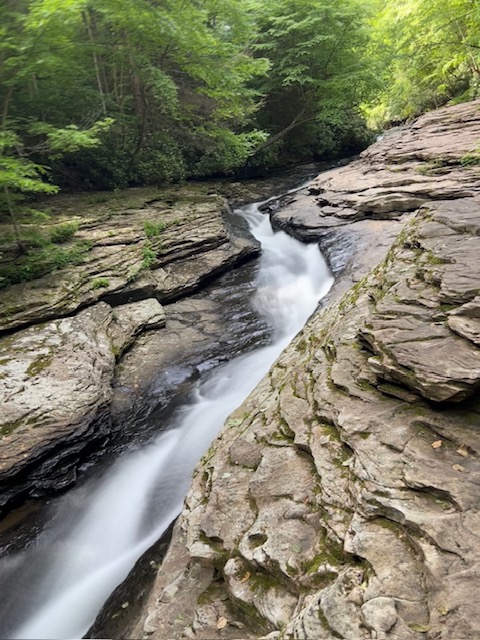 Long-exposure photograph of a natural waterslide cascading through smooth, layered rock formations in a lush green forest. The silky white water winds through a rocky channel surrounded by moss-covered stone and dense trees, creating a peaceful and secluded natural scene.