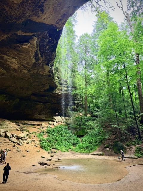Ash Cave in Hocking Hills State Park featuring a massive sandstone recess cave, seasonal waterfall, and lush green forest surrounding the amphitheater-like rock formation.
