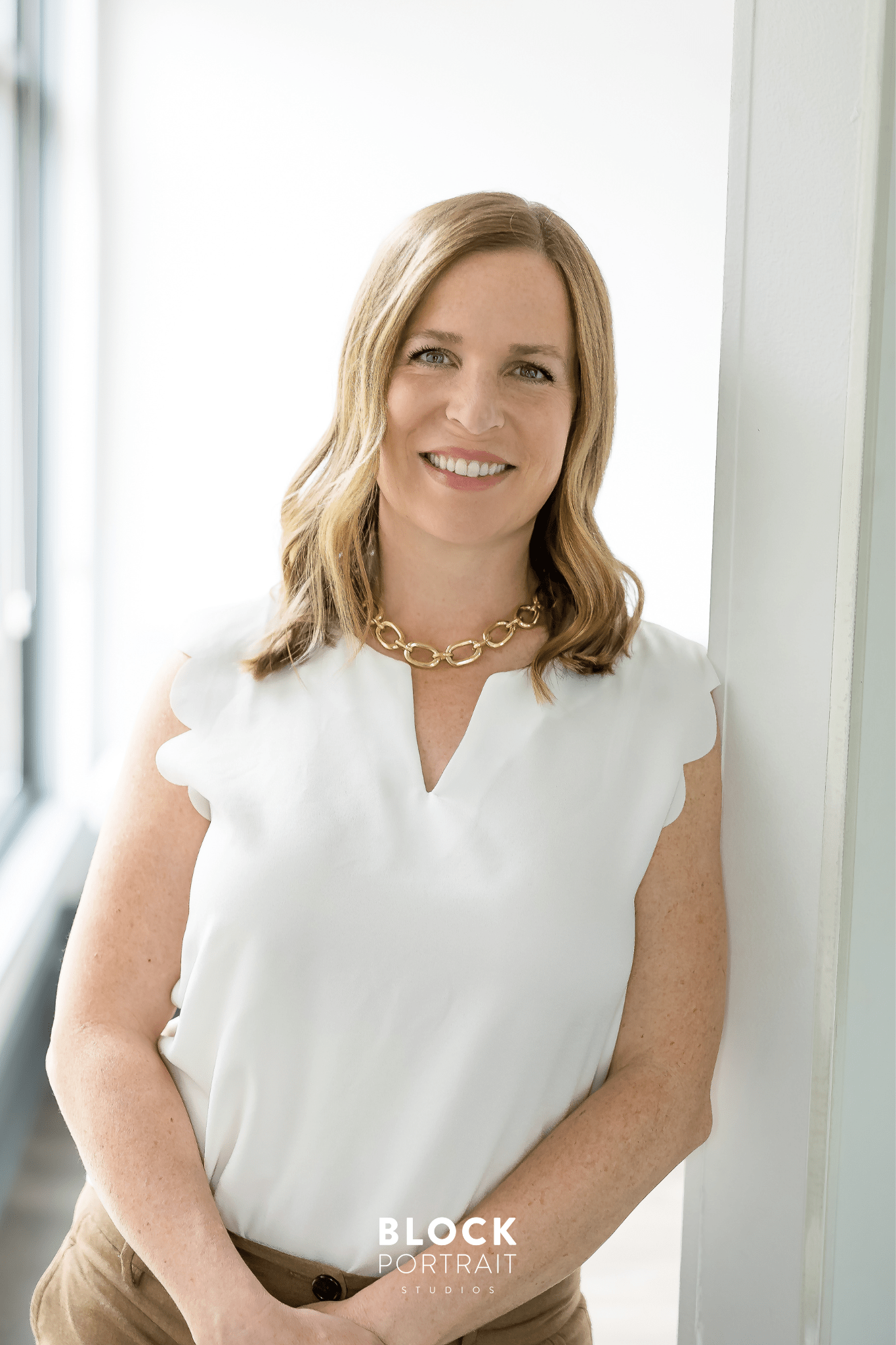 Professional headshot by Block Portrait Studios of a caucasian woman with blonde hair and blue eyes, wearing a white blouse with beige pants, standing and leaning against a white wall with windows behind her, while smiling directly at the camera.