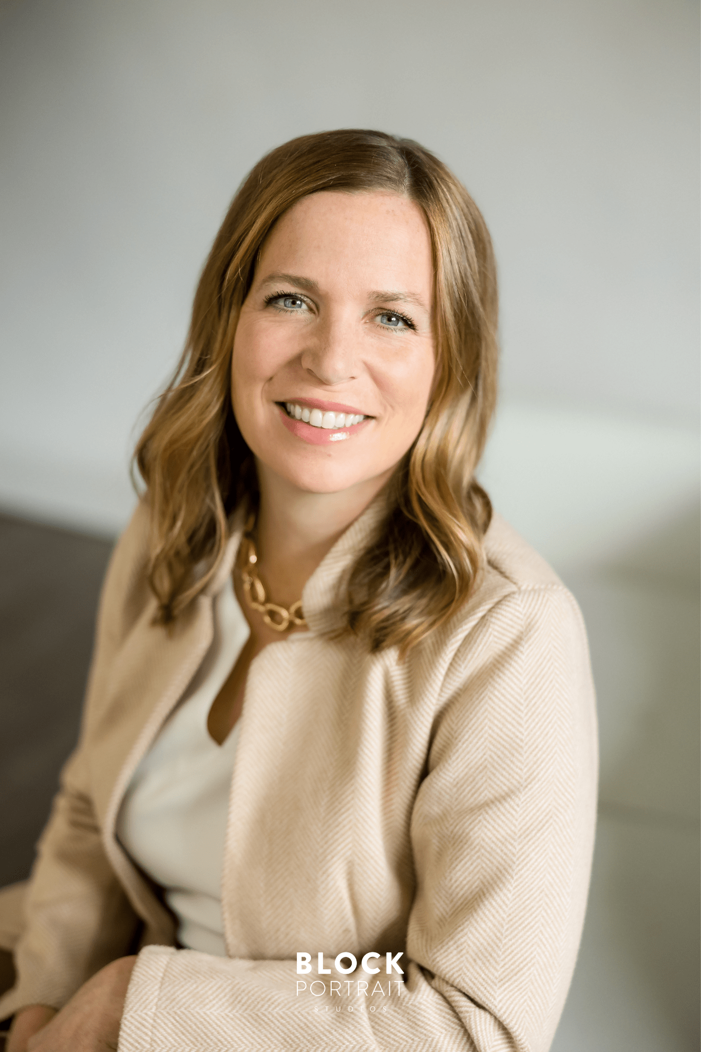 Headshot of a caucasian woman with blonde hair and blue eyes wearing a beige blazer and a white top, sitting on a white couch, smiling at the camera by Block Portrait Studios a photography studio that specializes in business branding.