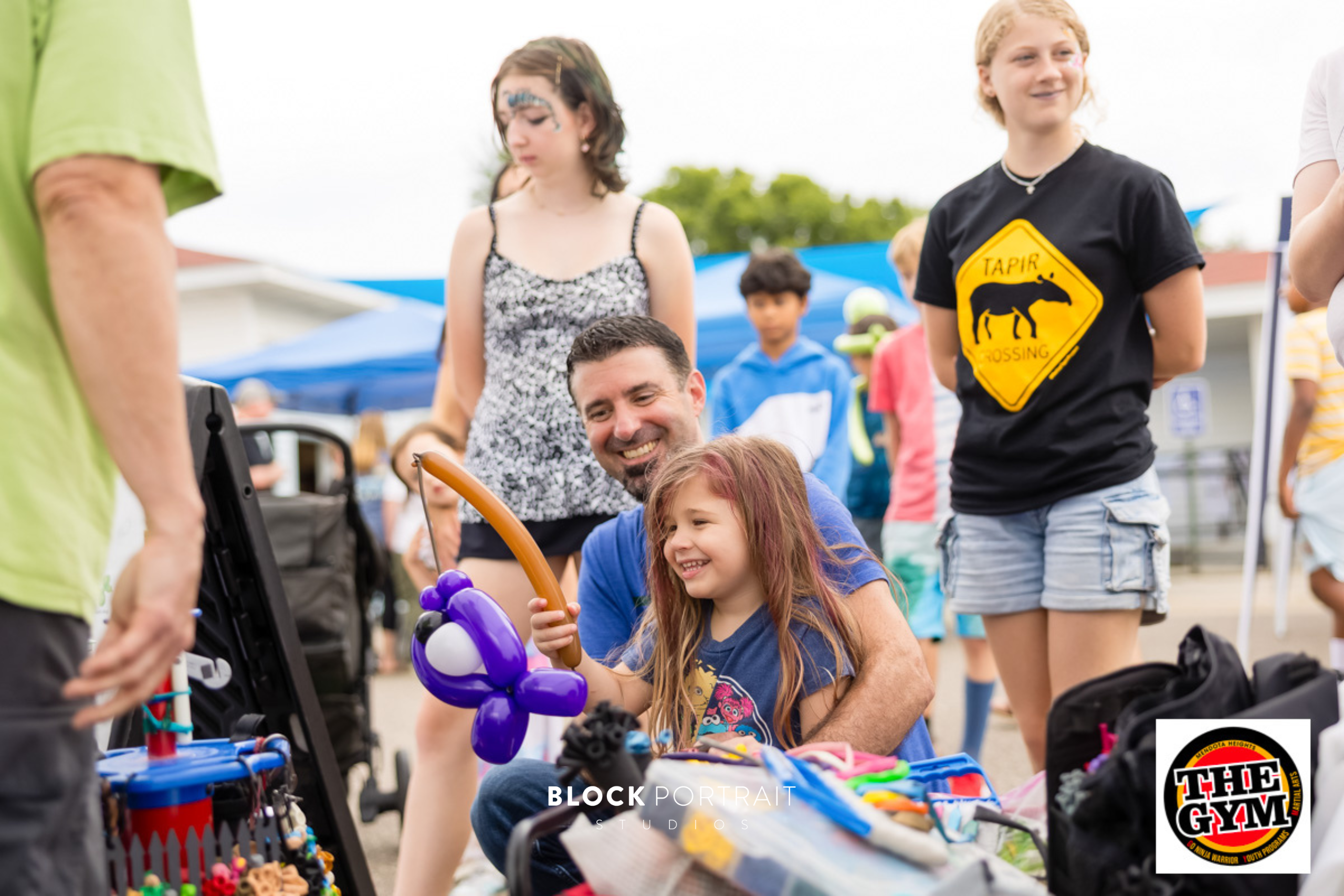 A dad smiling as his daughter receives balloon art shaped like a purple fish on a fishing rod.