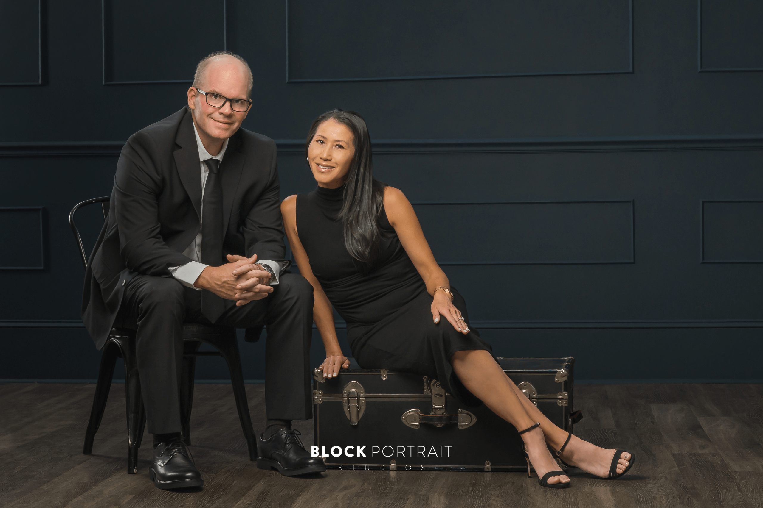 A woman sitting on a black luggage case wearing a black knee-length dress next to a man in a black suit sitting in a chair.