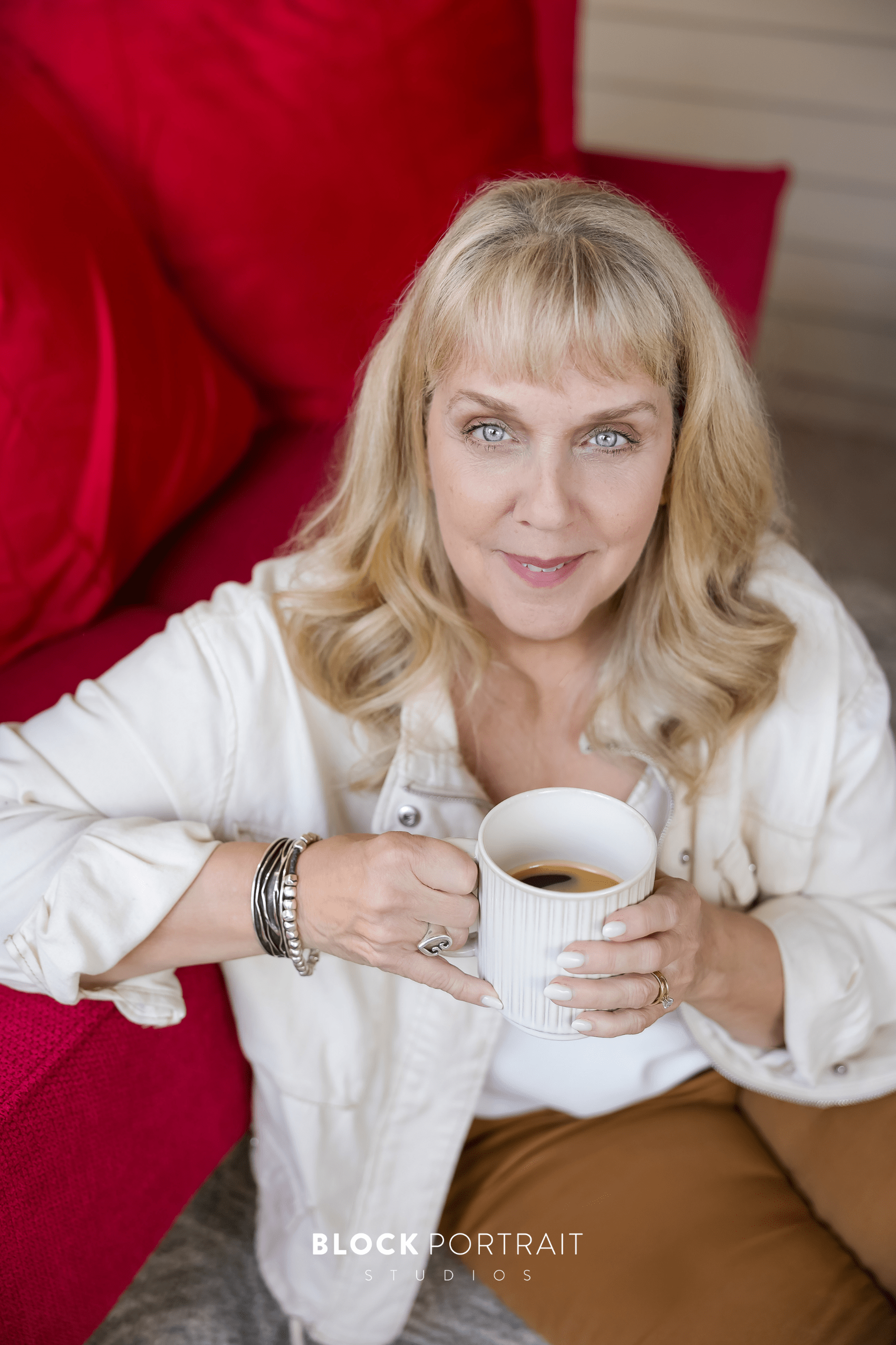 A woman sitting in front of a red couch holding a white cup of coffee and wearing a cream button-up and soft neutrals.