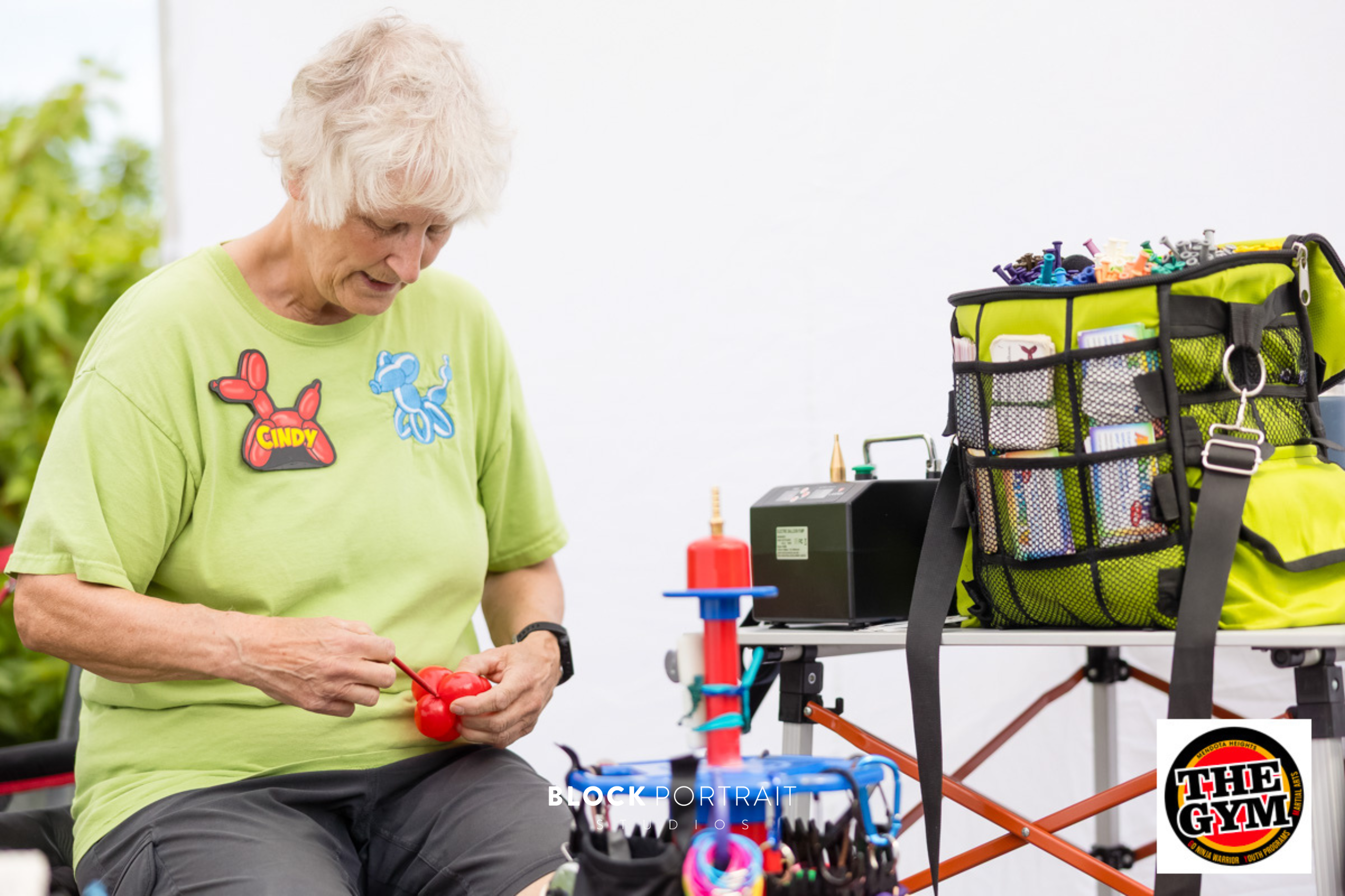 A woman in a green shirt with a balloon animal shaped name badge blowing up a red balloon.
