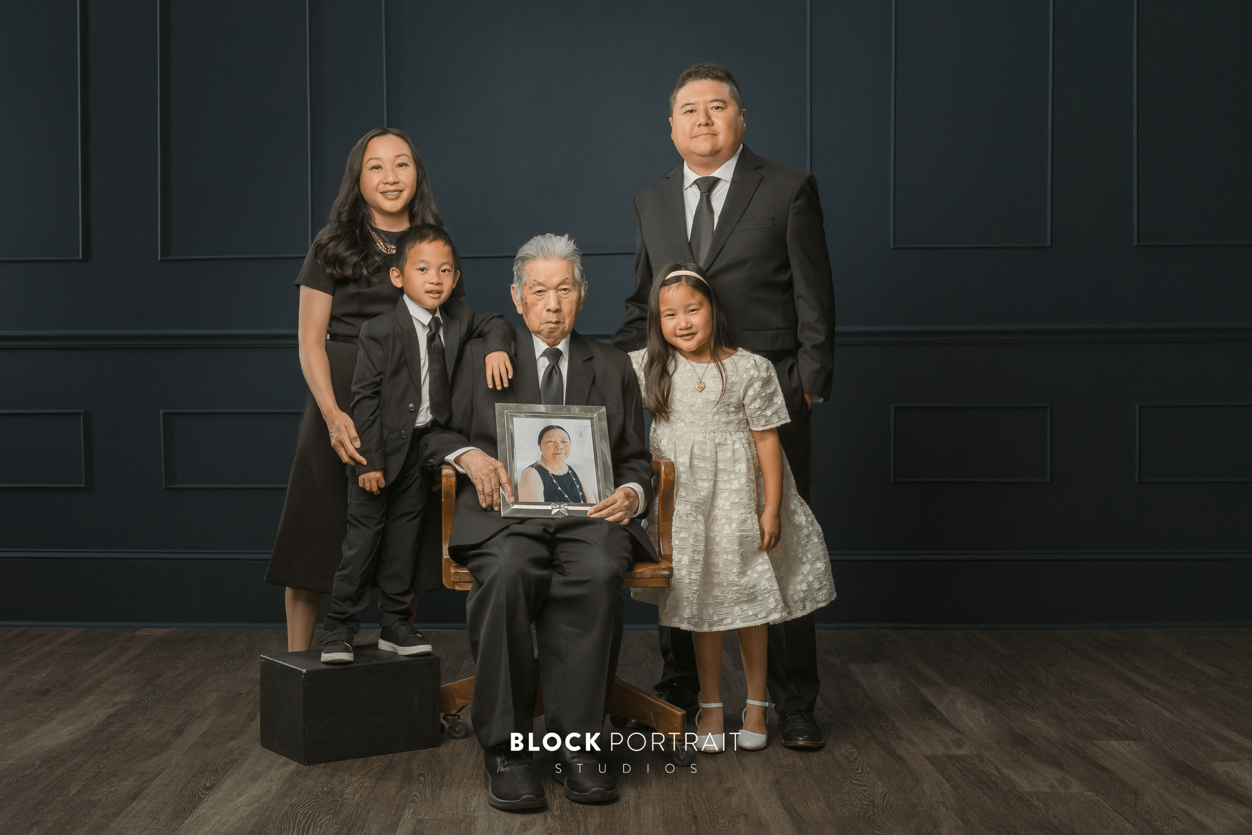 A family of four in formalwear standing around an elderly man sitting in a chair holding a framed photo of his late wife.