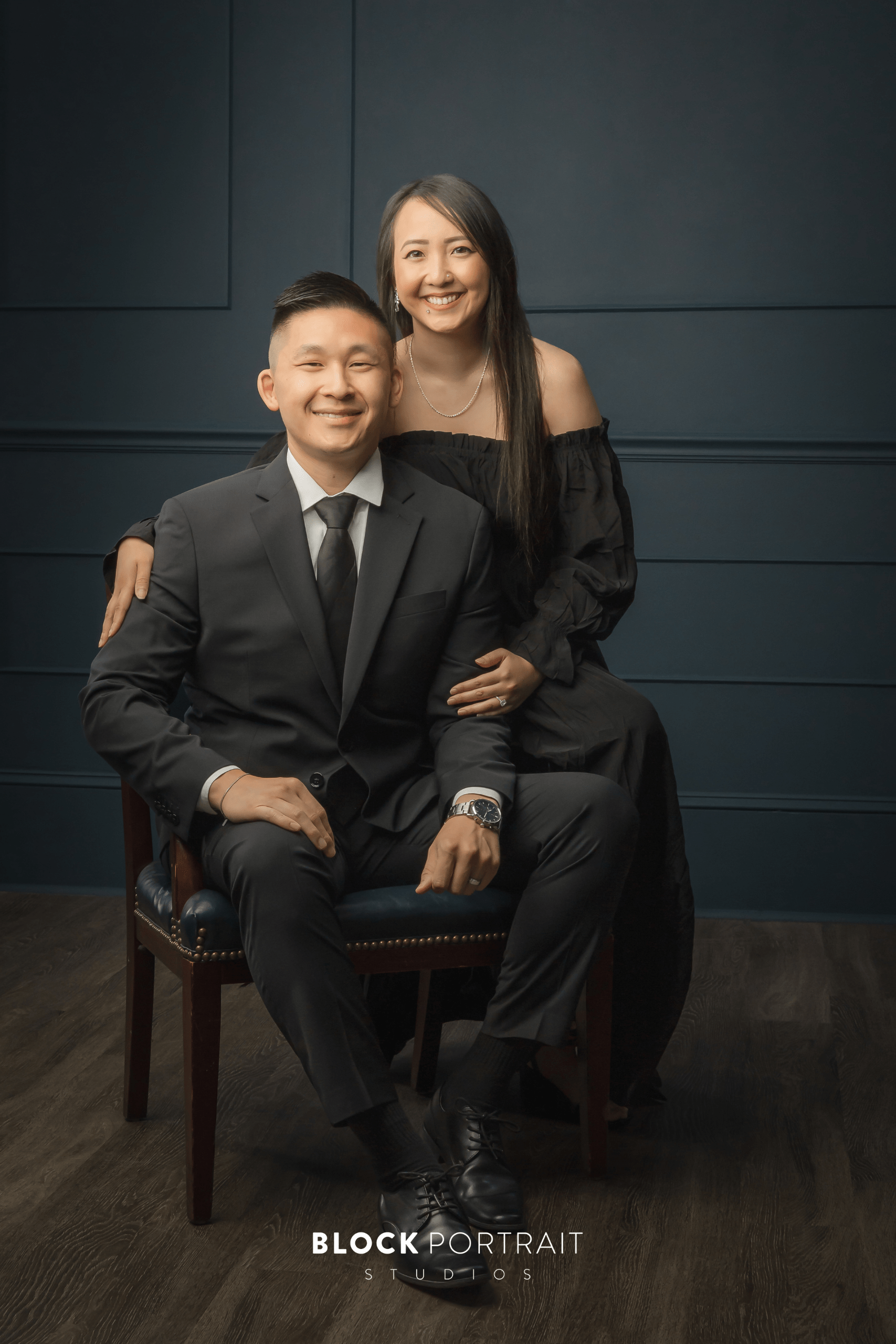 A man in a black suit sitting in a chair with a woman in a formal length black gown standing behind him with her arms wrapped around him.