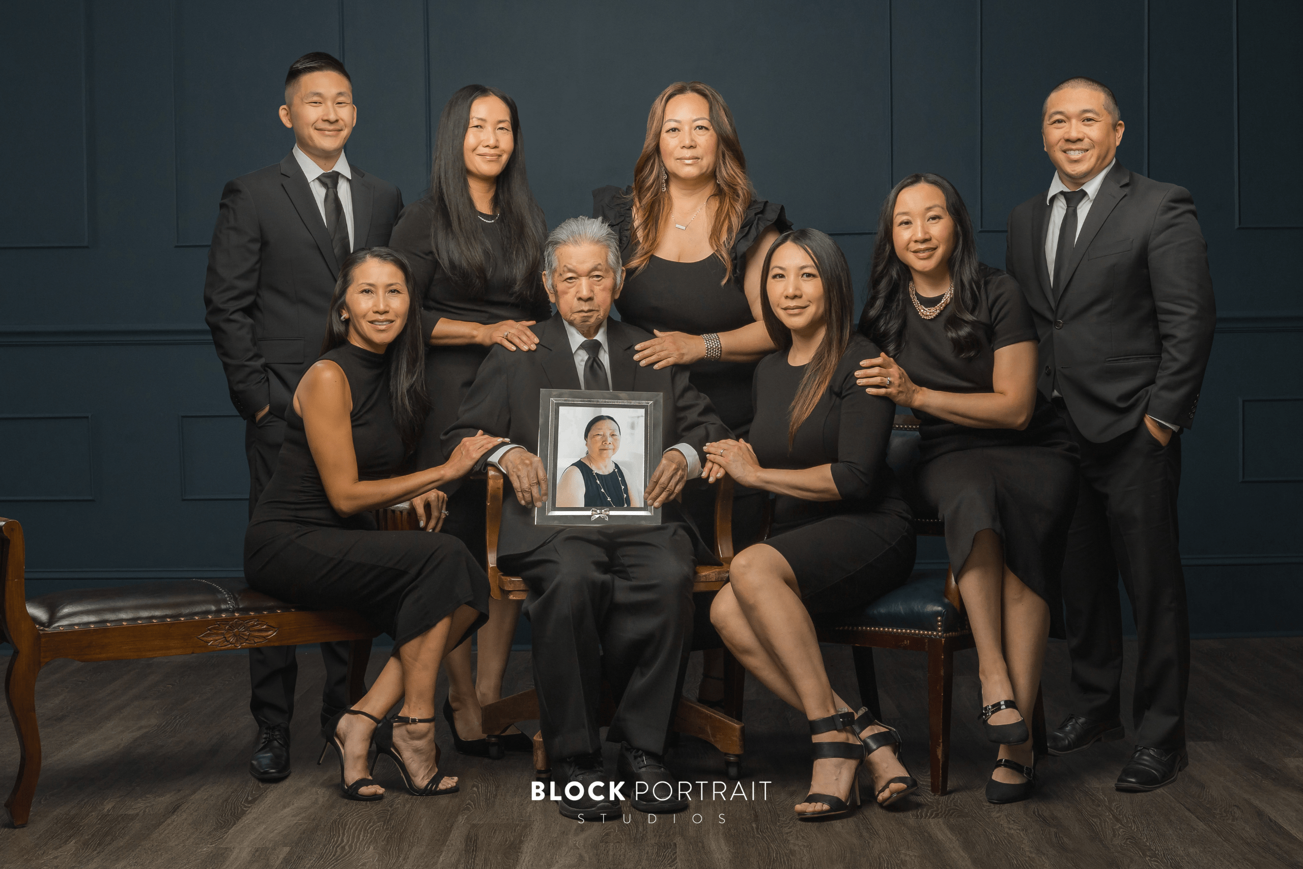 An elderly Asian man sitting in a chair holding a framed photo of his late wife and surrounded by his seven adult children all wearing black formalwear.
