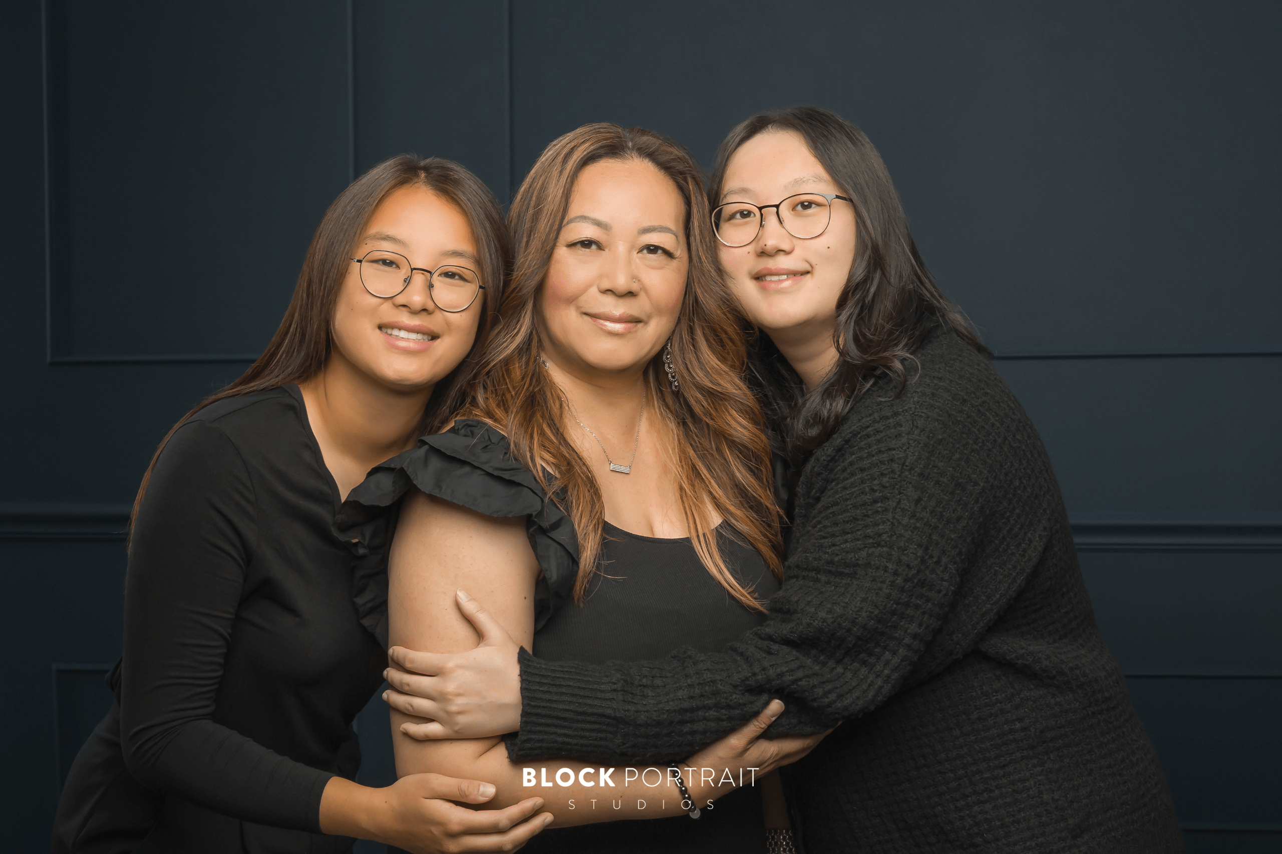 A mother and her two adult daughters all wearing black formalwear while hugging their mother.