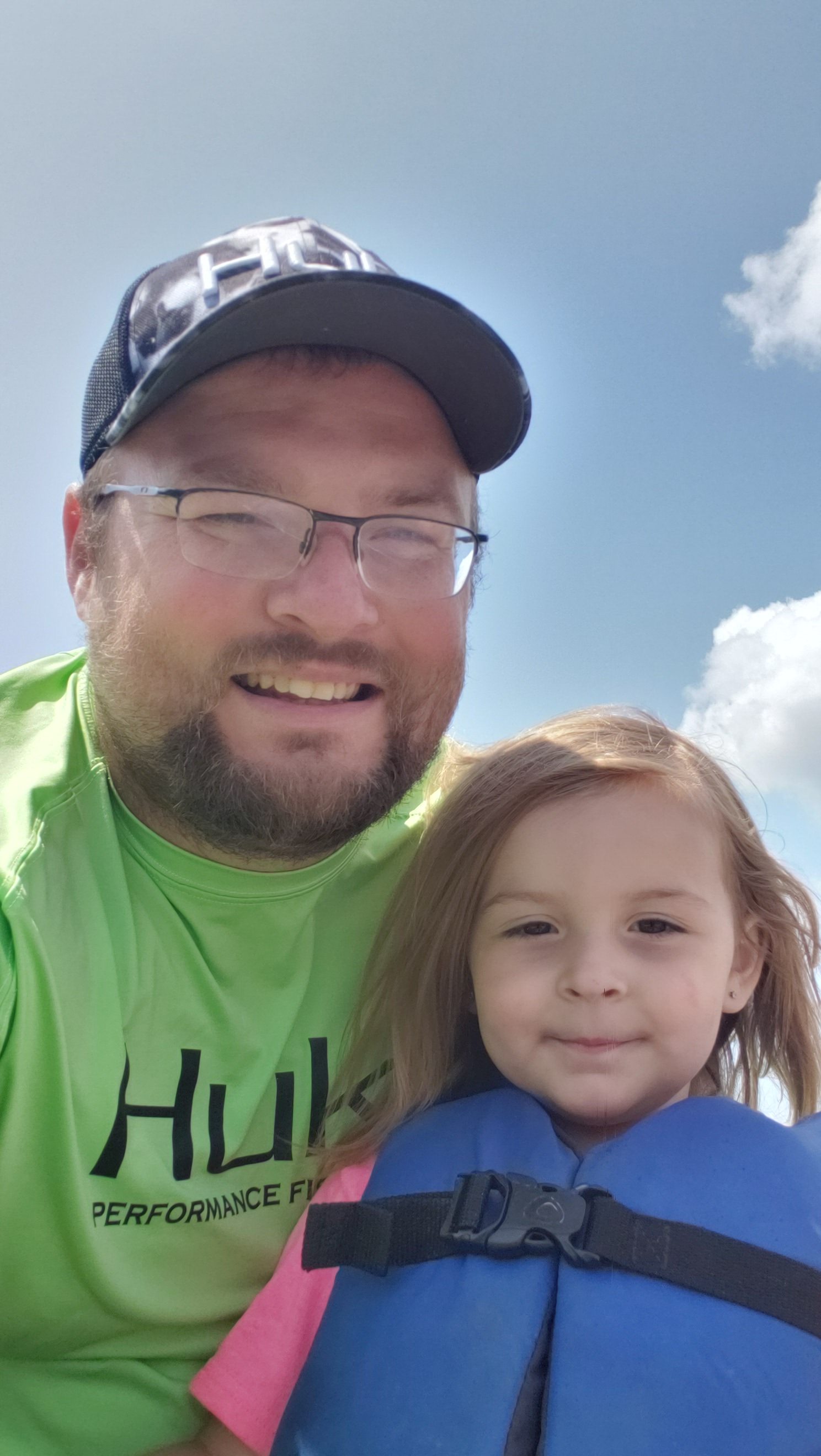 Brian McLucas and his daughter Addilyn smile outside. Addilyn is wearing a life vest and the blue sky is behind them.