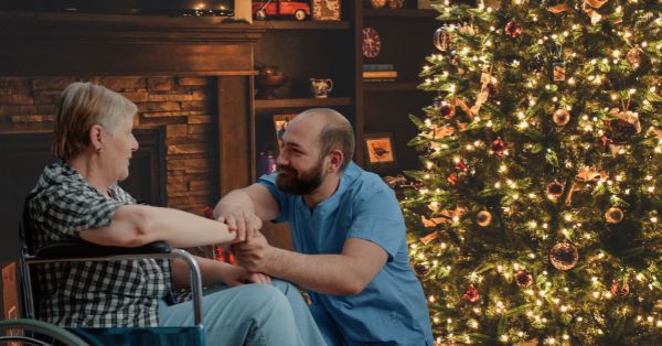 Photo of elderly woman sitting in a wheelchair in a living room decorated brightly for the holidays with a tree and lights. A younger man with a beard and balding head kneels smiling next to her and offering a supporting hand.