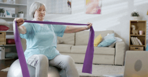 GIF: Short video of a woman with gray hair sitting on an exercise ball using a purple resistance band to stretch her arms. 