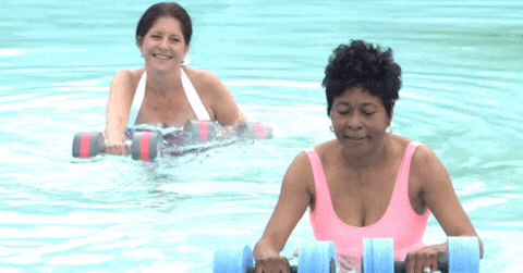 GIF: Short clip of a black and a caucasian elderly women doing water aerobics in a clean pool with floating weights.