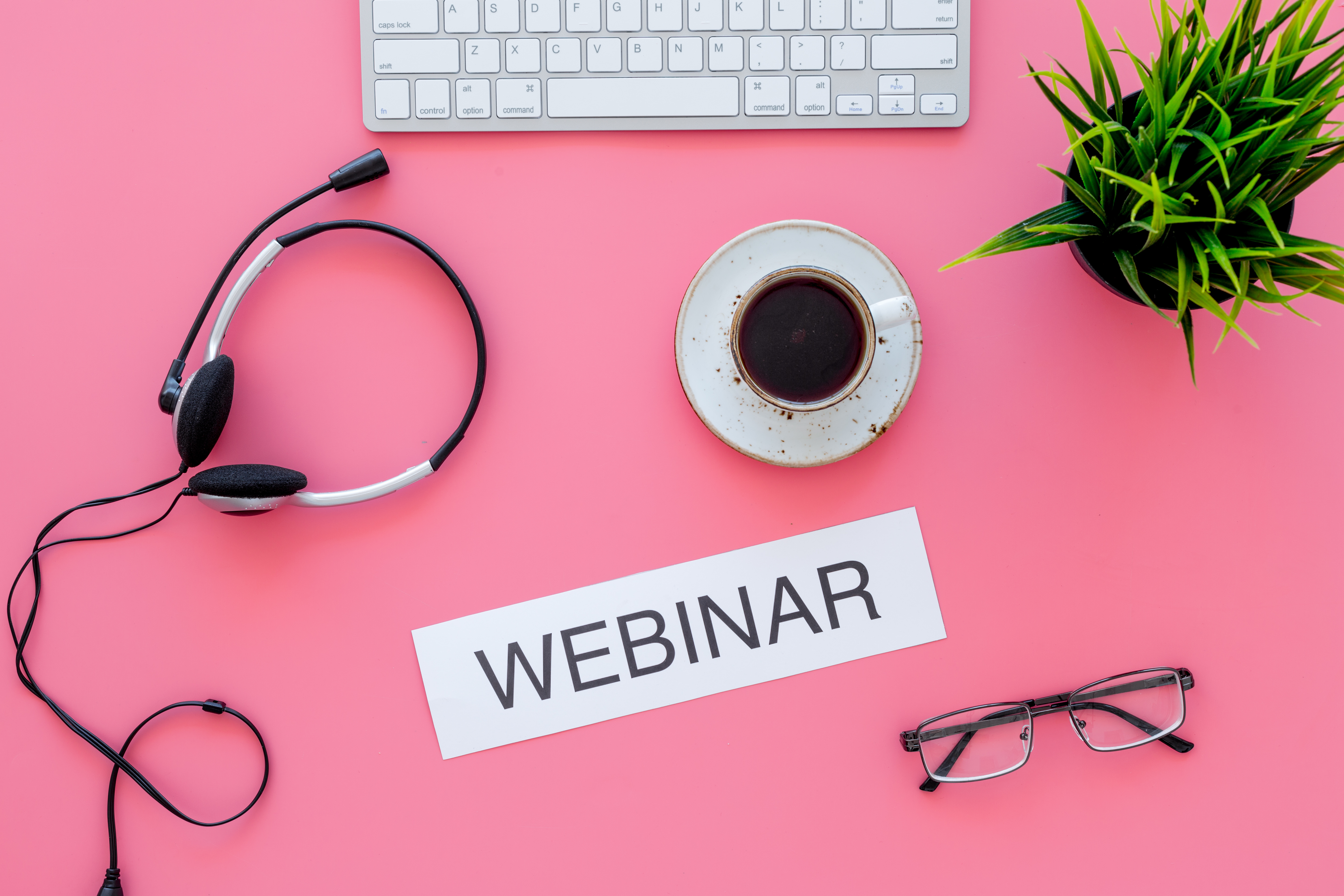 laptop, coffee, headphones, glasses and succulent plant on a pink background with the word "webinar" on a strip of paper