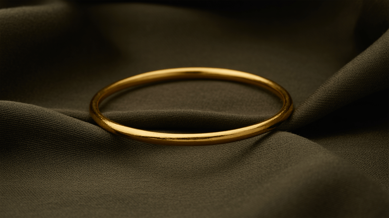 A plain 22 karat gold bangle resting on dark textured cloth under warm low side light
