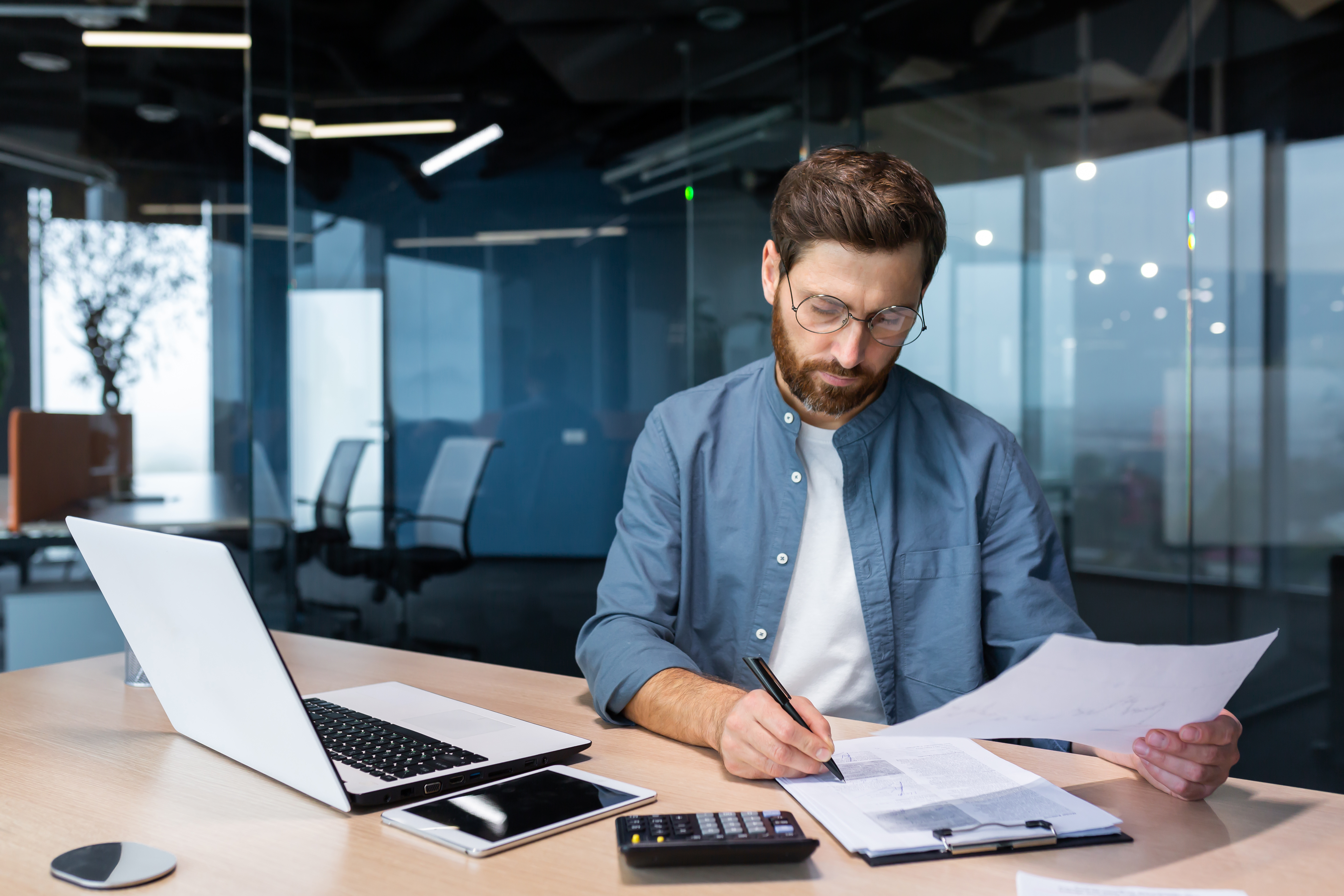 Serious and focused financier accountant on paper work inside office, mature man using calculator and laptop for calculating reports and summarizing accounts, businessman at work in casual clothes