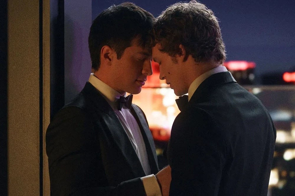 Dramatic shot of the two leads from the Heated Rivalry series in formal wear, touching foreheads against a city skyline background.