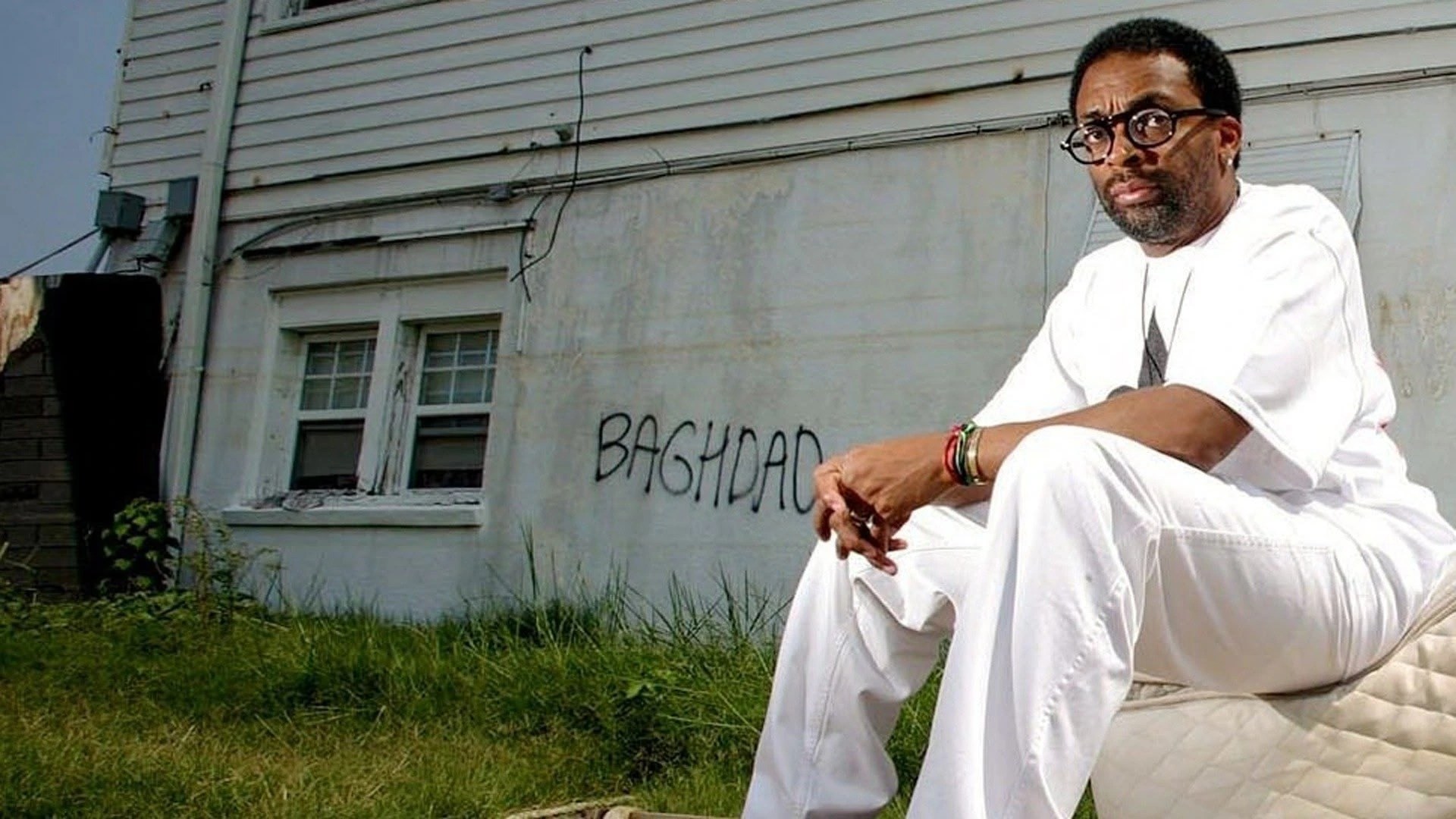 Spike Lee sitting outdoors in all-white clothing in front of a damaged building with "BAGHDAD" graffiti, representing the aftermath of Hurricane Katrina in New Orleans.
