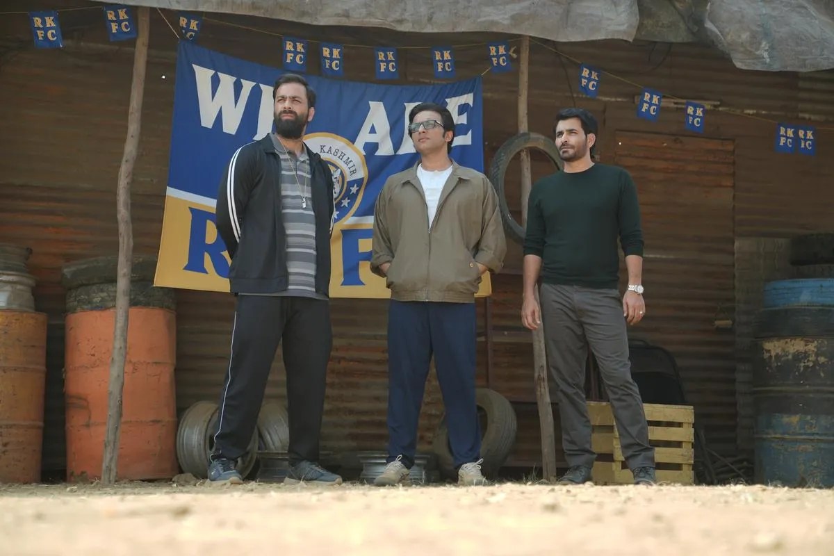 Three Real Kashmir Football Club team members standing proudly in front of a blue club banner on a rugged dirt pitch, capturing the series' authentic visual style.
