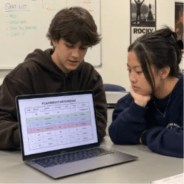 Two film students collaborating on a digital production schedule using software in a classroom setting.