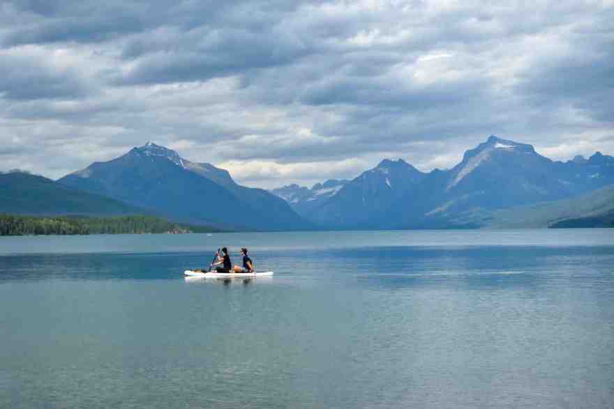 Two students on a paddle board on the lake at Glacier National Park with mountains in the background.. 