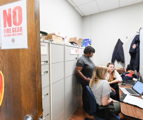 The students sit in an office while the director stands and all three look at their laptop. The door to the office reads, "Chattanooga Fire Department."