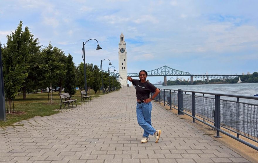 A person stands smiling on a wide paved walkway along a river, pointing toward a tall white clock tower in the distance. Trees and benches line one side of the path, while black street lamps and a metal railing run along the river’s edge. In the background, a large green truss bridge spans the river under a partly cloudy sky.