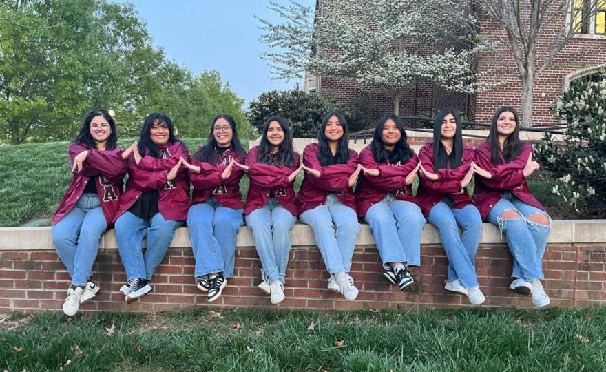 Eight women wearing matching maroon jackets and blue jeans sit side by side on a low brick wall outdoors, linking their arms to form an interlocking hand sign. Green grass and leafy trees surround them, and a brick building with blooming white-flowered trees is in the background.