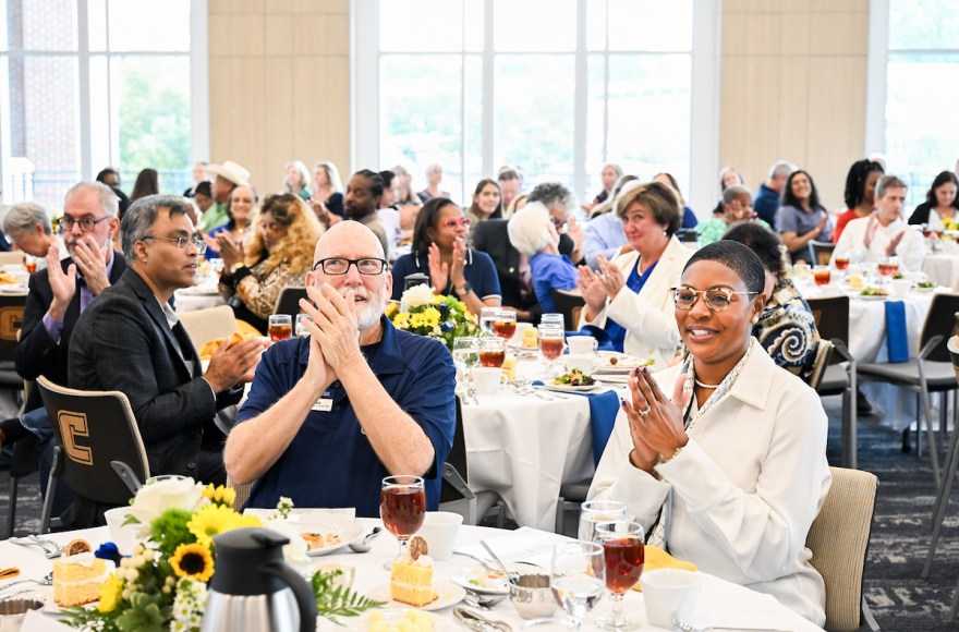 A large group of attendees seated at round tables clapping and smiling during the 2025 UTC Service Awards luncheon, with plates of food, iced tea, and floral centerpieces on the tables.