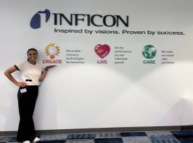 Student Hailey Henderson stands indoors, smiling and gesturing toward a colorful corporate wall display for INFICON with the words “Inspired by visions. Proven by success.” and icons labeled CREATE, LIVE and CARE with short taglines about technology, growth, and safety.