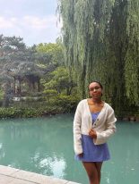 Student Hailey Henderson stands near a turquoise pond surrounded by lush greenery and trees, including a large weeping willow. Garden structures are partially visible in the background under a partly cloudy sky.