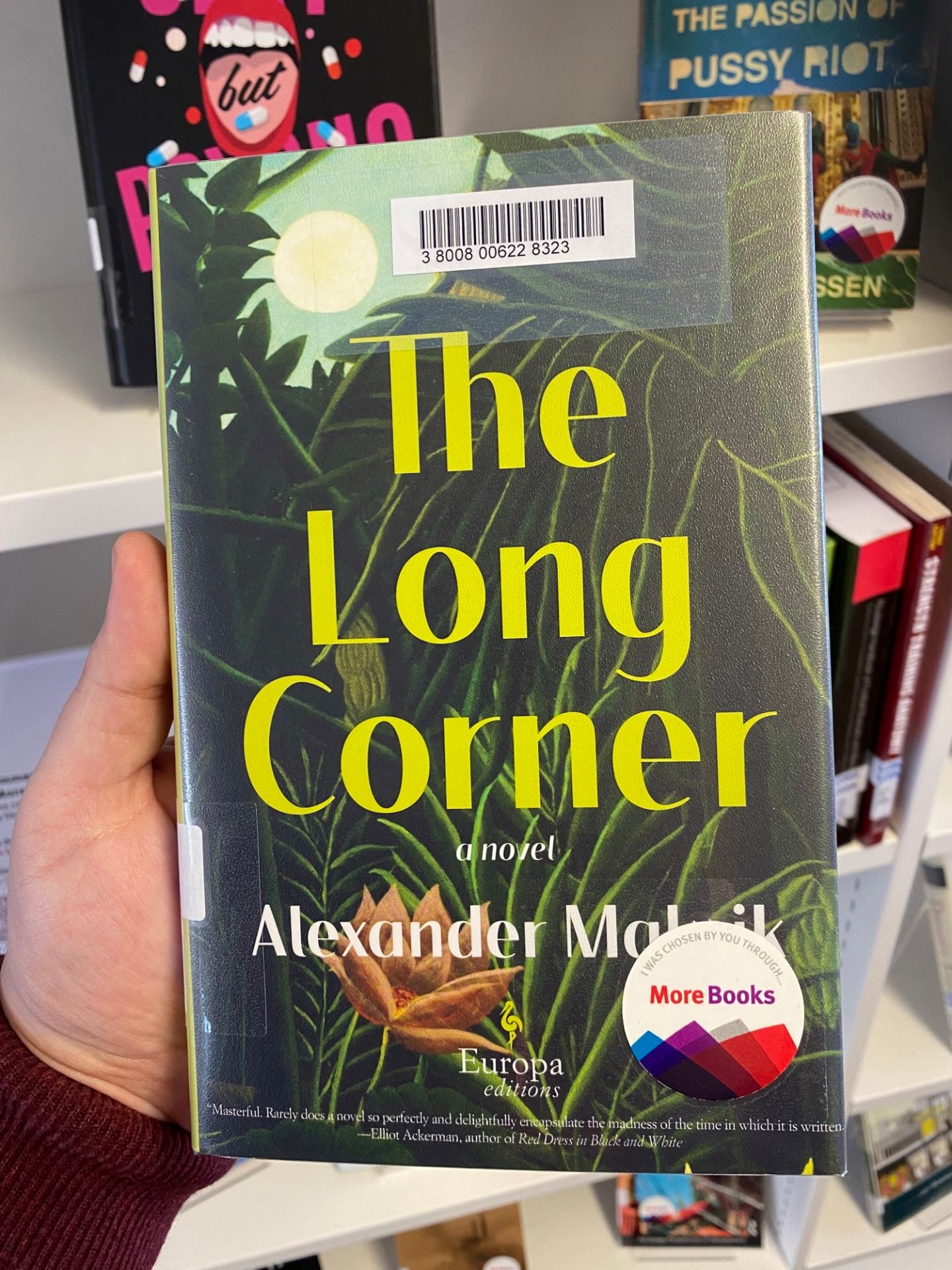 A hand is holding a book entitled The Long Corner, by Alexander Maksik. The book has a "More Books" sticker on the cover.
