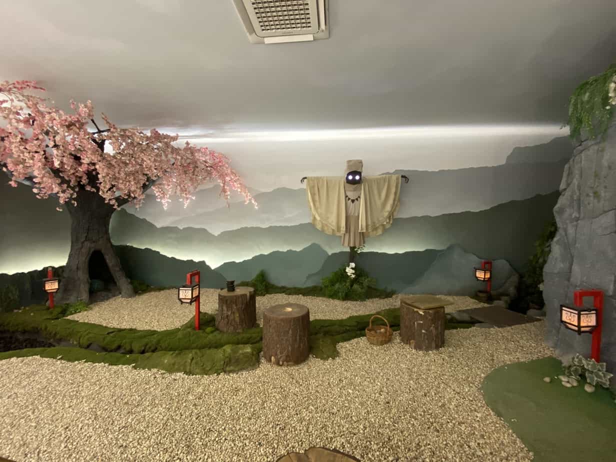 Kuebiko, the Crumbling Prince, who looks like a scarecrow standing in a Japanese stone Zen garden, behind a stream with moss covered banks. There are tree stumps, Japanese lanterns, a cherry blossom tree in full bloom and shoji screens. There are mountains in the distance. There is a basket in the garden.