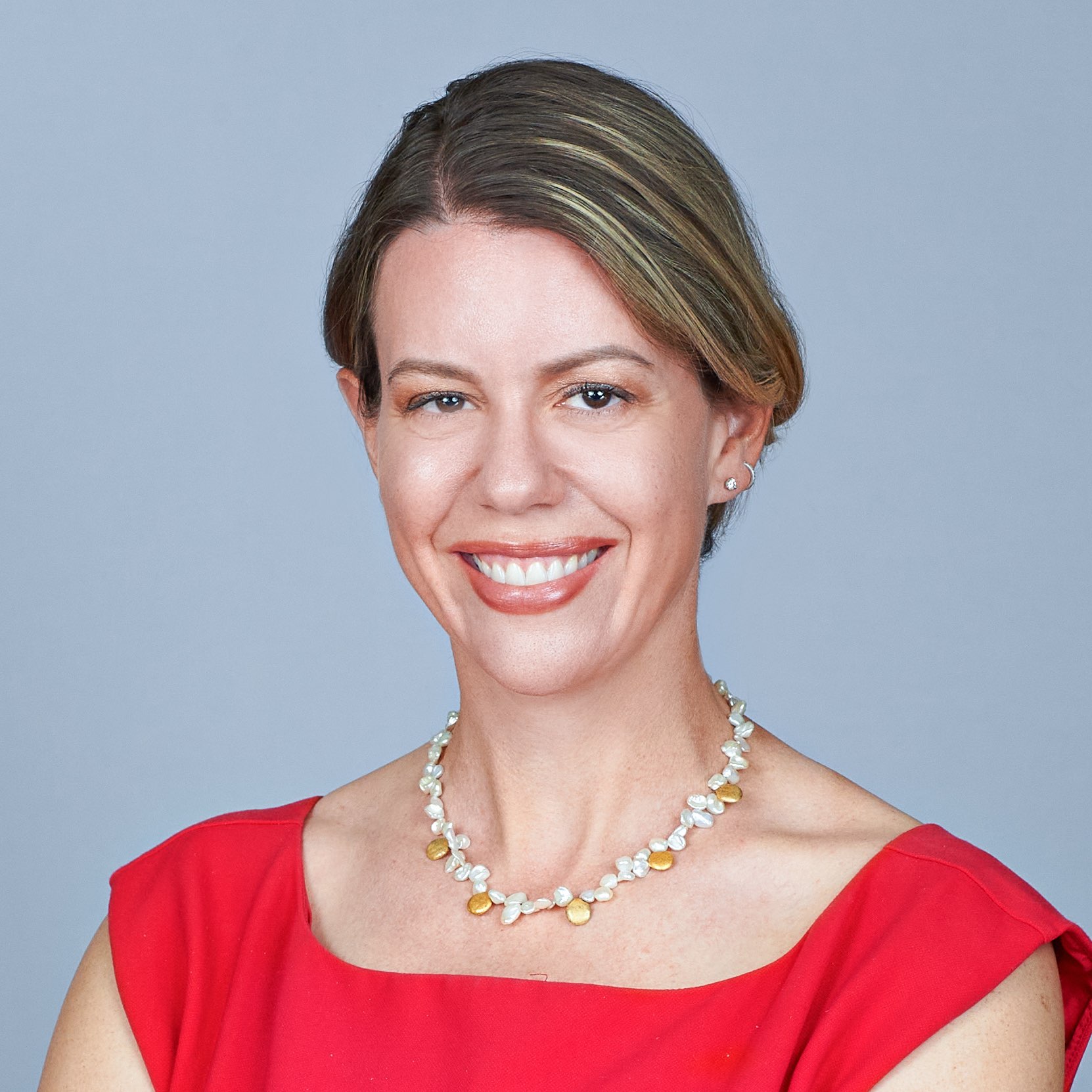 Portrait of Katie Pryal, a white woman with blond hair, smiling, wearing a pink shirt, standing on a green hill in front of palm trees.