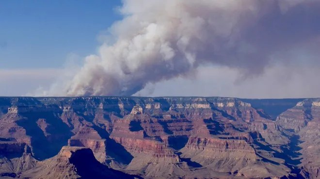 A plume of smoke rises over the Grand Canyon, indicating a wildfire in the north rim area, with striking rock formations visible in the foreground.
