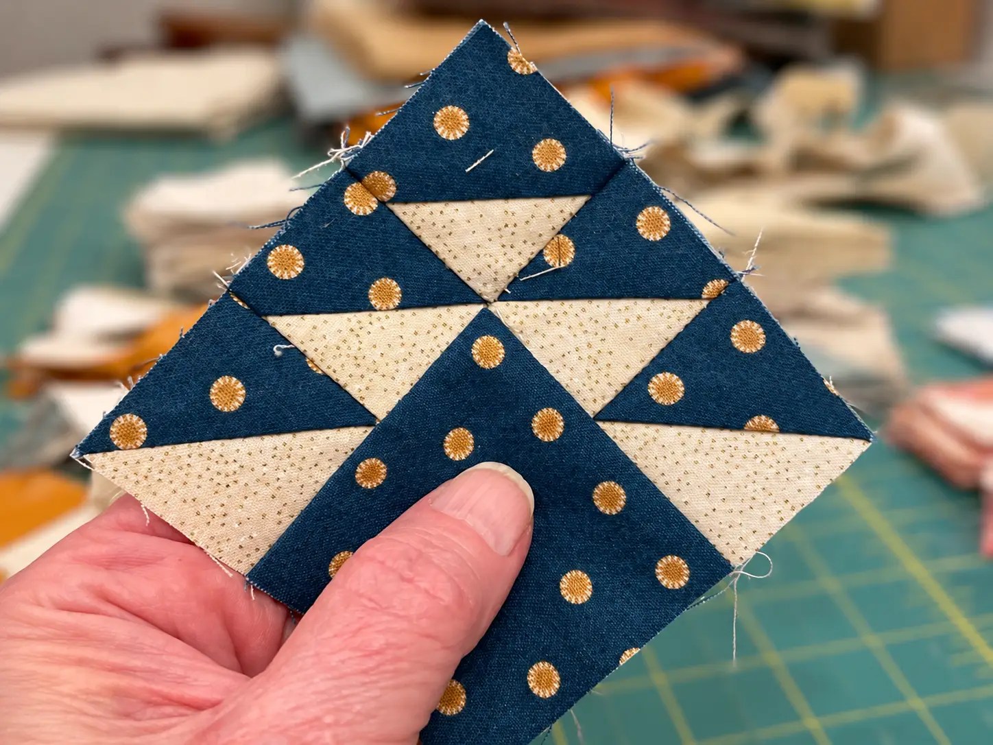 Hand holding a three inch flying geese quilt block made with navy fabric with gold dots and cream triangles above a green cutting mat.