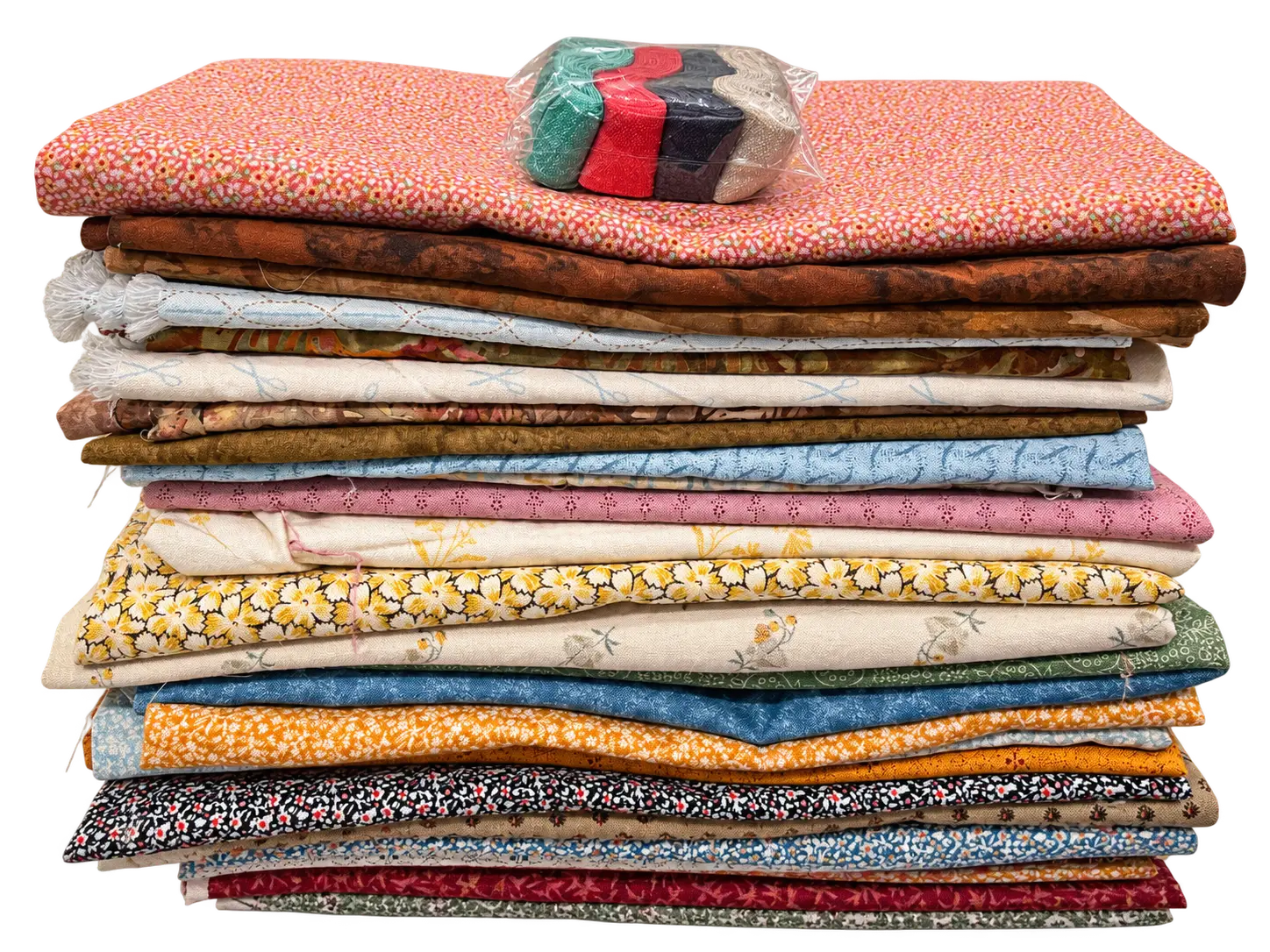 Stack of folded quilting fabrics in colorful floral and geometric prints, topped with a clear packet of teal, red, black, and beige trim.
