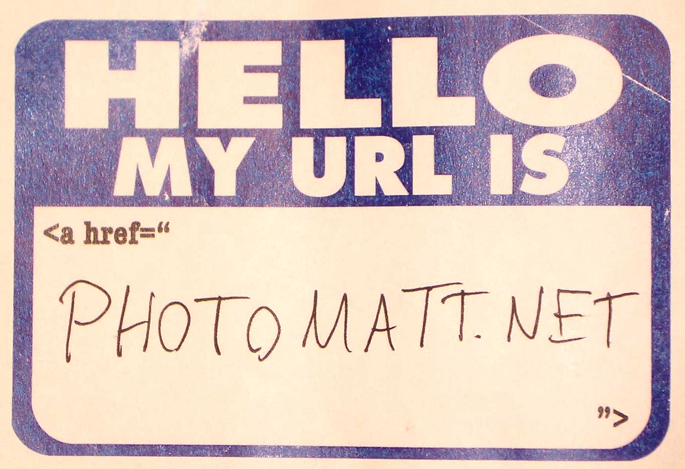 A common sticker-type name tag that is preprinted with the large text "Hello My URL is" with smaller text underneath that reads and a blank space in the middle where someone has handwritten in pen "photomatt.net"